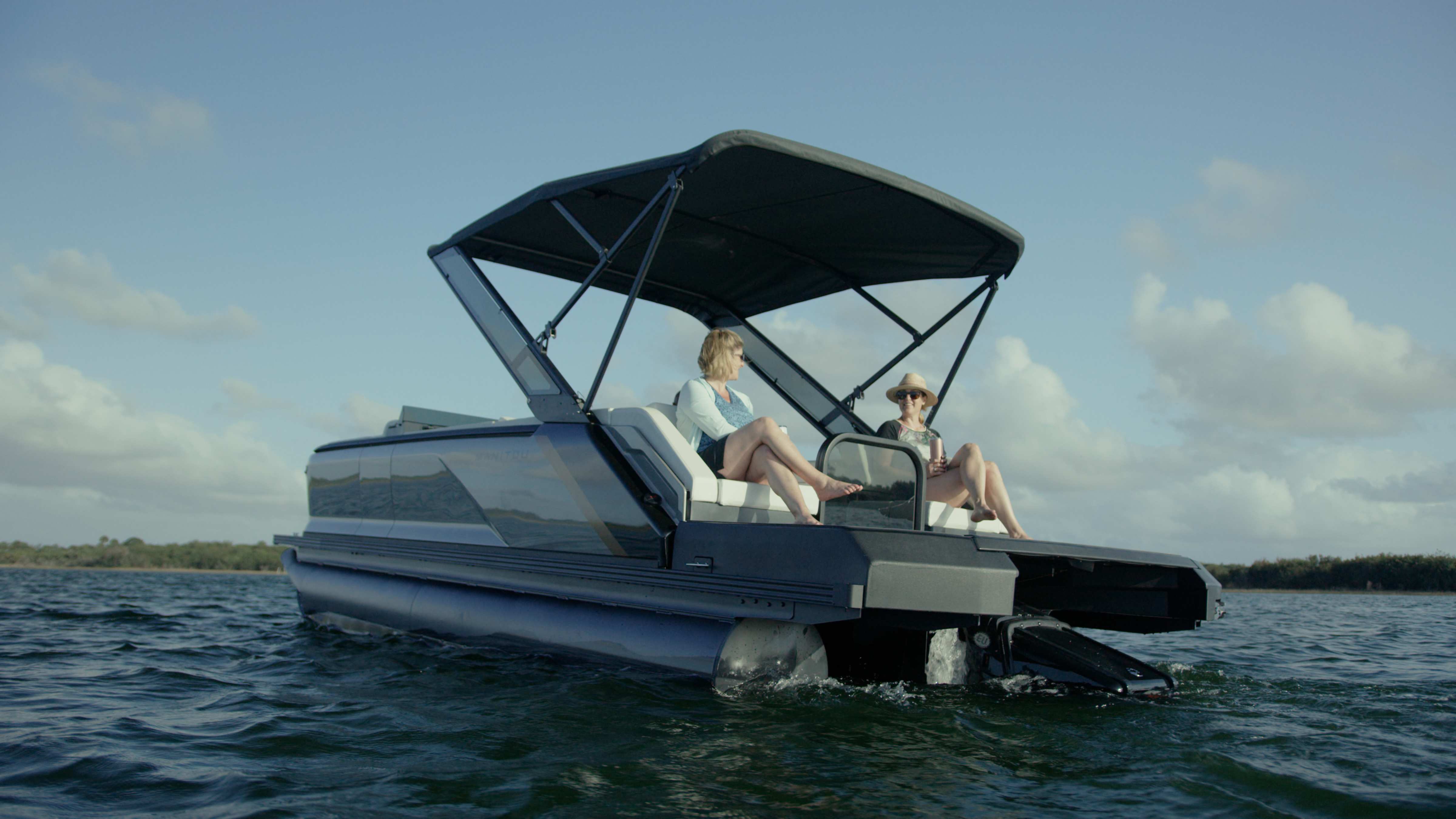 Two passengers talking on a 2023 Manitou Explore sport Pontoon Boat with a Bimini top